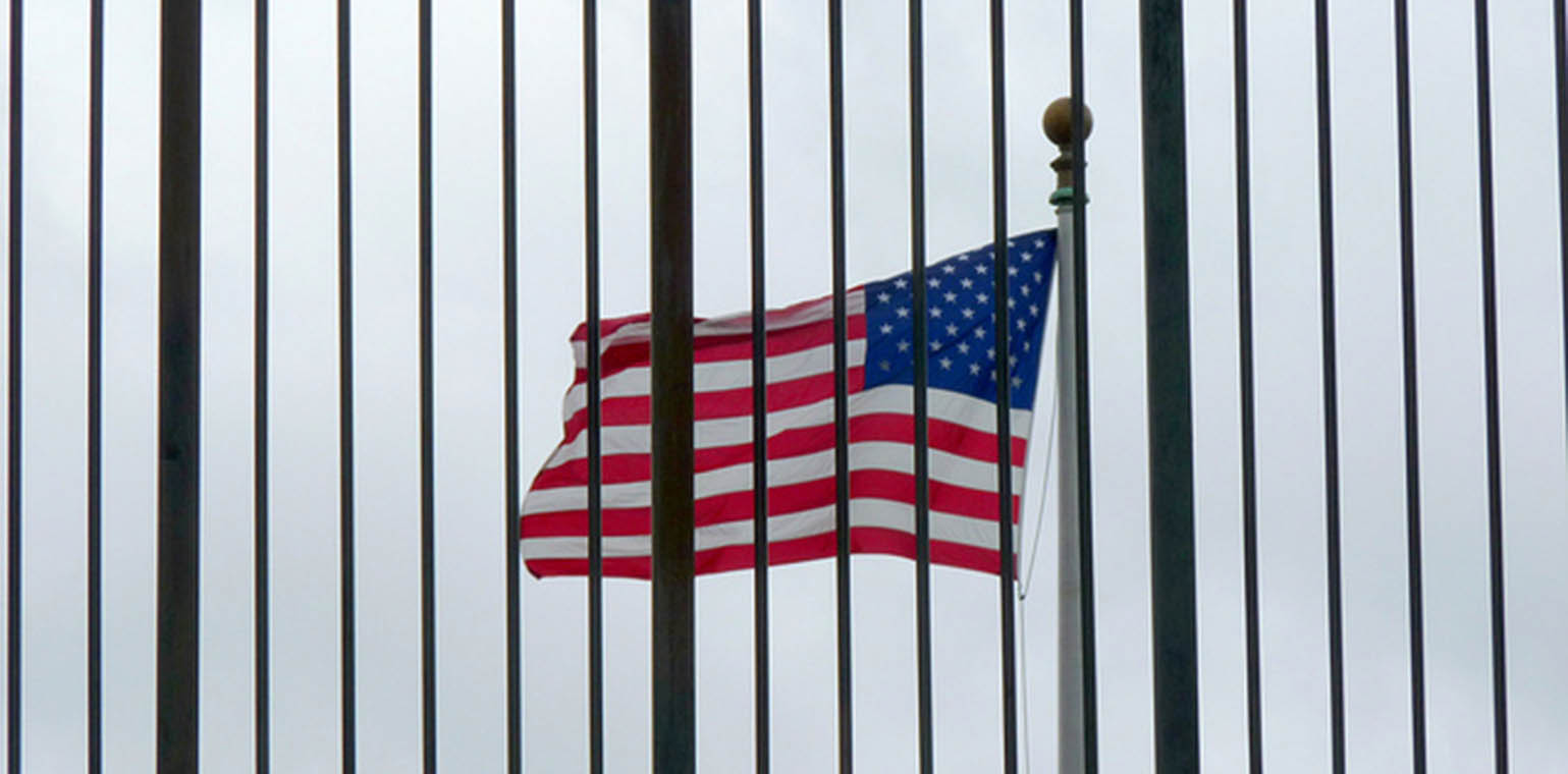 Photo of a U.S. flag flying toward the left, with iron bars of the U.S.-Mexico border fence in the foreground.
