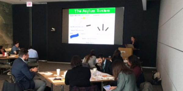 Photo of a conference room where individuals in suits sit at tables taking notes. At the front of the room is a projection screen showing a slide titled "The Asylum System"